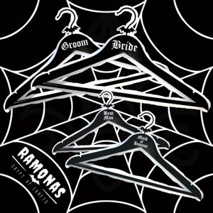 May include: Black wooden hangers with white lettering reading "Groom", "Bride", "Best Man", and "Maid of Honour". Each hanger has a bat-shaped hook. The background features a white spiderweb pattern on a black surface.