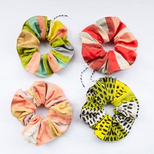 May include: Four patterned scrunchies in a variety of colours. One is striped red and white, another is green, yellow, and peach. The third is peach and white, and the fourth is yellow with black dots. Each has a black and white string.