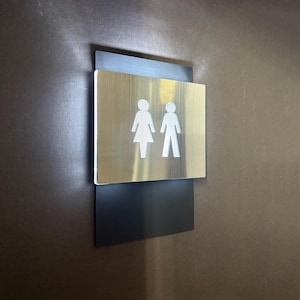 May include: A restroom sign with a brushed metal finish. The sign features a square shape with a black rectangular backing. The sign displays the silhouettes of a woman and a man.
