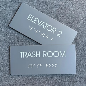 Op de afbeelding: Twee rechthoekige grijze bordjes met witte letters en braille. Het bovenste bordje vermeldt ELEVATOR 2 en het onderste bordje vermeldt TRASH ROOM. Beide bordjes hebben een gladde, matte afwerking met duidelijke tekst en verhoogde braillepunten.