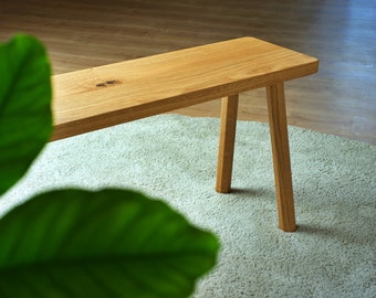 May include: A light brown wooden bench featuring a smooth, rectangular seat with a visible natural wood grain pattern and a small dark knot. The bench is supported by four sturdy, slightly angled legs made from the same light-toned wood material.