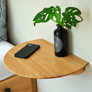 Könnte beinhalten: Ein halbrunder Holzregal an einer Wand montiert, mit einem schwarzen Telefon und einer schwarzen Vase mit einer grünen Pflanze darauf.