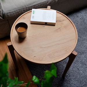 Könnte beinhalten: Ein runder Holz-Beistelltisch mit einer hellen Holzoptik. Der Tisch hat drei Beine und eine Tasse Kaffee sowie ein Buch darauf. Das Buch trägt den Titel "Sapiens" von Yuval Noah Harari.