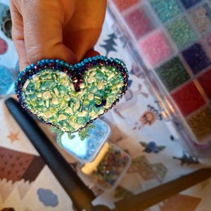May include: A heart-shaped beaded brooch in shades of green, blue, and white. The brooch is intricately detailed with small beads, creating a textured surface. The edges are outlined with darker blue and purple beads. The brooch is held up by a hand.