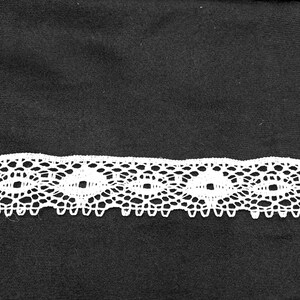 May include: A length of white lace with a repeating openwork design, set against a black backdrop. The lace pattern includes interconnected oval shapes and scalloped edges. Ideal for crafting, this lace offers a delicate and decorative element to projects.