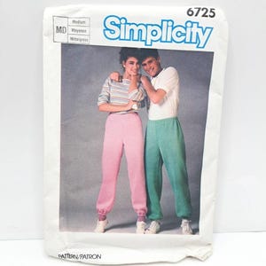 May include: Simplicity pattern envelope with a vintage image of a couple in athletic pants. The woman wears pink trousers and a striped top, the man wears green trousers and a white top. The envelope is white with blue text.