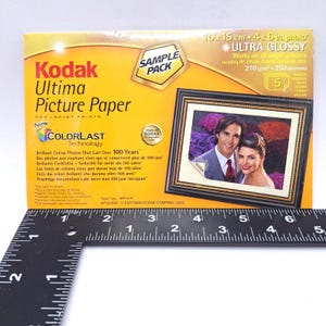 Op de afbeelding: Een gele doos Kodak Ultima fotopapier voor inkjetprinters. De doos heeft een ingelijste foto van een stel. Tekst op de doos is onder meer "ULTRA GLOSSY" en "Brilliant Colour Photos That Last Over 100 Years." De foto is 10 x 15 cm groot.
