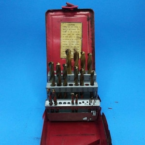 Op de afbeelding: Een vintage rode metalen boorbitsset met een scharnierend deksel. De set bevat meerdere boorbits van verschillende maten, gerangschikt in een metalen houder. De boorbits zijn goudkleurig en de behuizing heeft een label met tekst. De achtergrond is blauw.