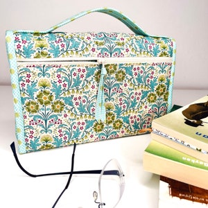Pode incluir: Uma bolsa para livros com estampa floral com al&ccedil;a, fecho de z&iacute;per e borla. A bolsa apresenta um padr&atilde;o de flores e folhas verdes, azuis e vermelhas em um fundo branco. Um par de &oacute;culos e uma pilha de livros est&atilde;o em primeiro plano.