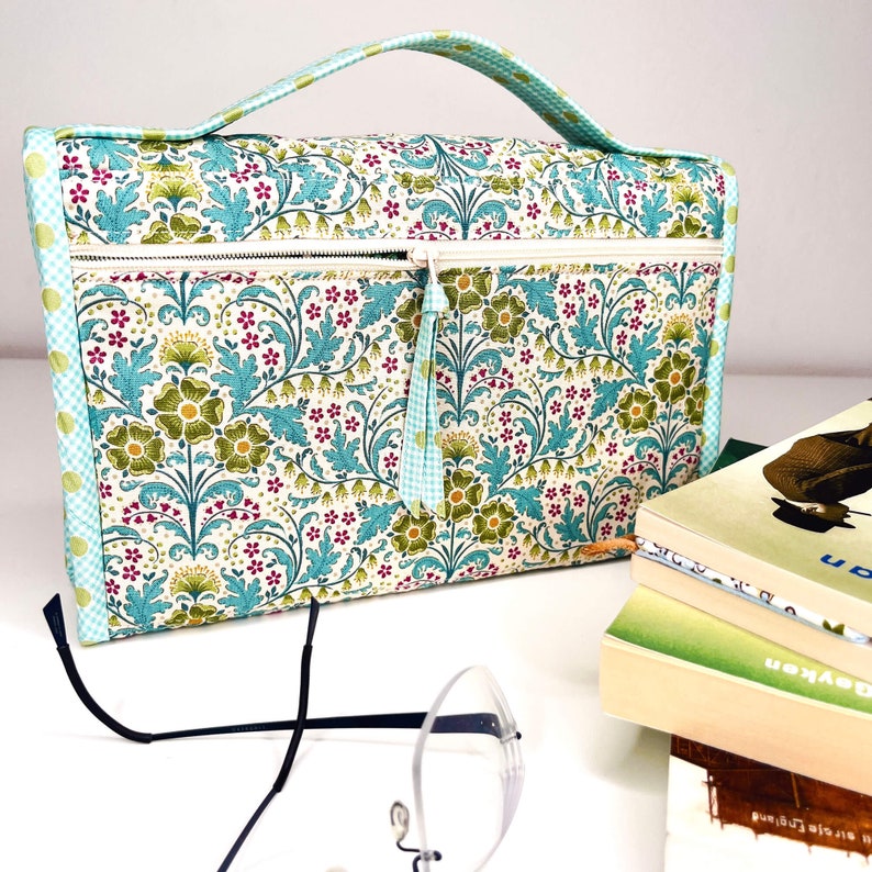 Pode incluir: Uma bolsa para livros com estampa floral com al&ccedil;a, fecho de z&iacute;per e borla. A bolsa apresenta um padr&atilde;o de flores e folhas verdes, azuis e vermelhas em um fundo branco. Um par de &oacute;culos e uma pilha de livros est&atilde;o em primeiro plano.