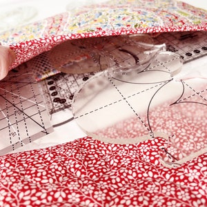 Puede incluir: Primer plano de una tela floral roja y blanca con una plantilla de pl&aacute;stico transparente para coser. La plantilla tiene forma de coraz&oacute;n y un patr&oacute;n de cuadr&iacute;cula.