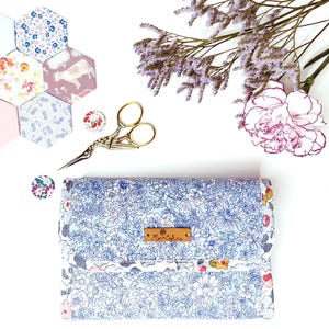 May include: A blue and white floral fabric needle minder with a brown leather label that says "The Little Stitcher". The needle minder is surrounded by fabric scraps, a pair of gold scissors, and a pink carnation.