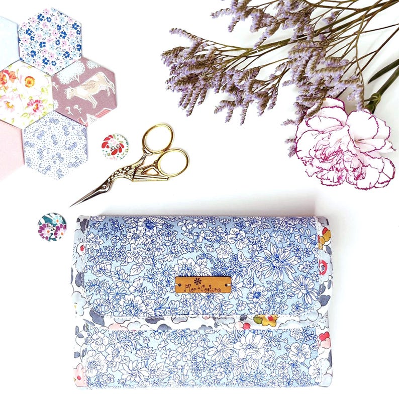 May include: A blue and white floral fabric needle minder with a brown leather label that says "The Little Stitcher". The needle minder is surrounded by fabric scraps, a pair of gold scissors, and a pink carnation.