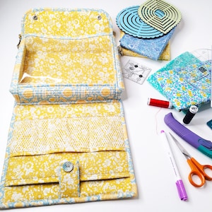 May include: A yellow floral fabric zippered pouch with a clear plastic window. The pouch is open and has multiple compartments inside. The pouch is surrounded by various sewing supplies, including fabric scraps, rulers, and a pair of scissors.
