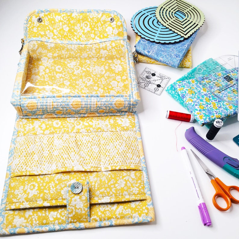 May include: A yellow floral fabric zippered pouch with a clear plastic window. The pouch is open and has multiple compartments inside. The pouch is surrounded by various sewing supplies, including fabric scraps, rulers, and a pair of scissors.