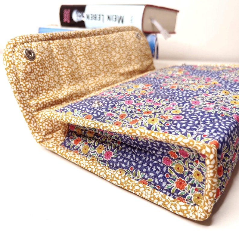 Pode incluir: Uma capa de livro acolchoada com um padr&atilde;o floral em tons de rosa, laranja e amarelo sobre um fundo azul marinho. A capa &eacute; adornada com tecido dourado e tem um fecho de press&atilde;o. A capa aberta revela o interior.