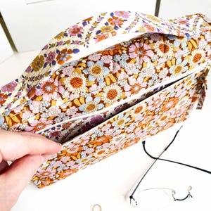 Pode incluir: Uma mala com estampa floral e al&ccedil;a. A mala apresenta um padr&atilde;o floral colorido em tons de laranja, rosa, amarelo e azul sobre um fundo marrom. O interior tem um z&iacute;per branco e um bolso. A al&ccedil;a &eacute; creme com um padr&atilde;o floral.