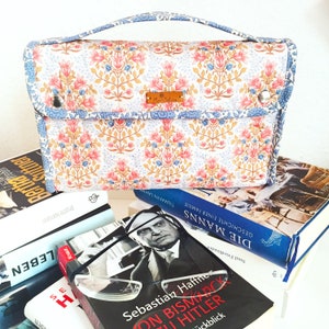 Pode incluir: Uma bolsa estampada com flores e uma al&ccedil;a, com flores rosa e azuis sobre um fundo branco. A bolsa est&aacute; colocada em cima de uma pilha de livros. Uma pequena etiqueta castanha est&aacute; presa &agrave; bolsa.