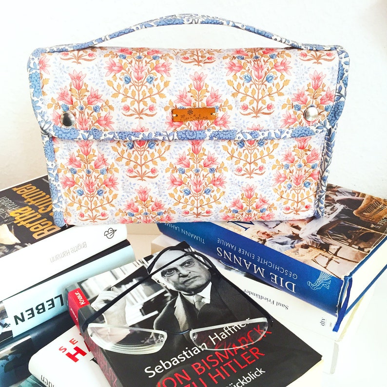 Pode incluir: Uma bolsa estampada com flores e uma al&ccedil;a, com flores rosa e azuis sobre um fundo branco. A bolsa est&aacute; colocada em cima de uma pilha de livros. Uma pequena etiqueta castanha est&aacute; presa &agrave; bolsa.