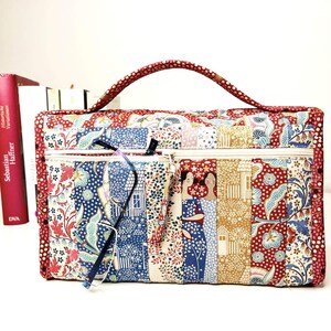 Pode incluir: Bolsa de tecido acolchoado com al&ccedil;a vermelha e fecho de z&iacute;per. A bolsa apresenta um design de patchwork com estampas florais e geom&eacute;tricas em vermelho, azul e creme. Um par de &oacute;culos est&aacute; no bolso com z&iacute;per. A bolsa tem uma ilustra&ccedil;&atilde;o decorativa de duas mulheres.