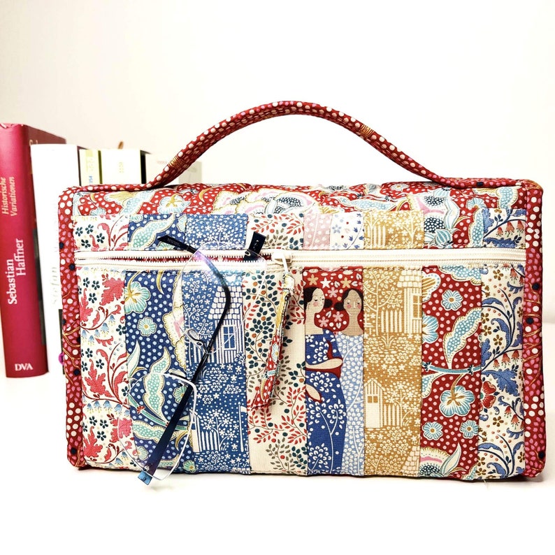 Pode incluir: Bolsa de tecido acolchoado com al&ccedil;a vermelha e fecho de z&iacute;per. A bolsa apresenta um design de patchwork com estampas florais e geom&eacute;tricas em vermelho, azul e creme. Um par de &oacute;culos est&aacute; no bolso com z&iacute;per. A bolsa tem uma ilustra&ccedil;&atilde;o decorativa de duas mulheres.