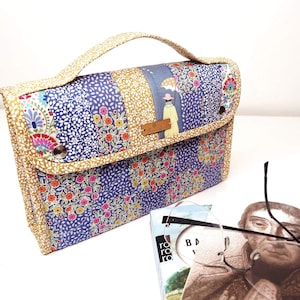 Pode incluir: Bolsa de m&atilde;o patchwork com um padr&atilde;o floral e de folhas em azul, dourado e rosa. A bolsa tem uma al&ccedil;a e um fecho de aba com um bot&atilde;o. Uma pequena etiqueta de madeira est&aacute; anexada. Um livro e &oacute;culos est&atilde;o em primeiro plano.