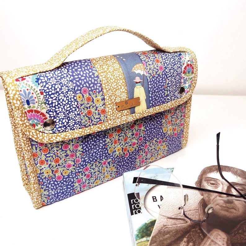 Pode incluir: Bolsa de m&atilde;o patchwork com um padr&atilde;o floral e de folhas em azul, dourado e rosa. A bolsa tem uma al&ccedil;a e um fecho de aba com um bot&atilde;o. Uma pequena etiqueta de madeira est&aacute; anexada. Um livro e &oacute;culos est&atilde;o em primeiro plano.