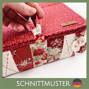 May include: A red and floral patterned fabric box with a button closure. The box features a patchwork design with red and white floral and polka dot fabrics. A small tag with embroidered details hangs from the side. The text "SCHNITTMUSTER" is at the bottom.