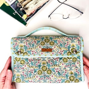 Pode incluir: Uma clutch com padr&atilde;o floral e al&ccedil;a, com fundo creme com flores e folhas verdes e azul-turquesa. A clutch tem uma borda xadrez vichy azul claro e uma pequena etiqueta de couro. A al&ccedil;a &eacute; azul claro.