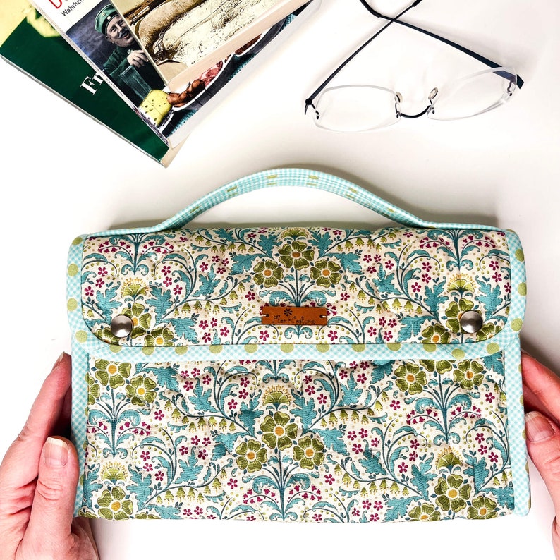 Pode incluir: Uma clutch com padr&atilde;o floral e al&ccedil;a, com fundo creme com flores e folhas verdes e azul-turquesa. A clutch tem uma borda xadrez vichy azul claro e uma pequena etiqueta de couro. A al&ccedil;a &eacute; azul claro.