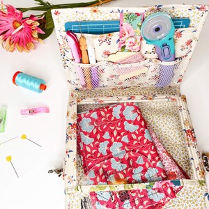 May include: A white fabric storage box with multiple pockets and compartments. The box is filled with various sewing supplies, including scissors, rulers, fabric scraps, and a rotary cutter. The box is open and the contents are visible.