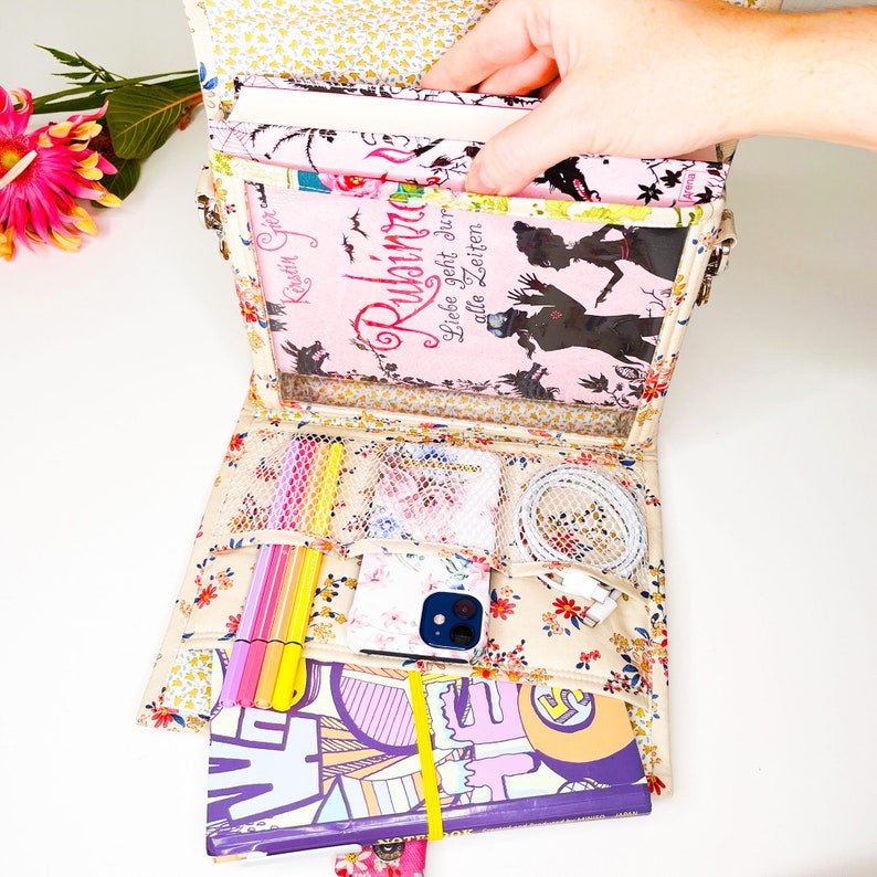 May include: A beige fabric bag with a clear plastic window, showing a stack of books with the title "Ruhimina Liebe geht durch alle Zeiten" and a floral pattern. The bag has multiple pockets and compartments, including a pocket for a phone and a pocket for a charger.