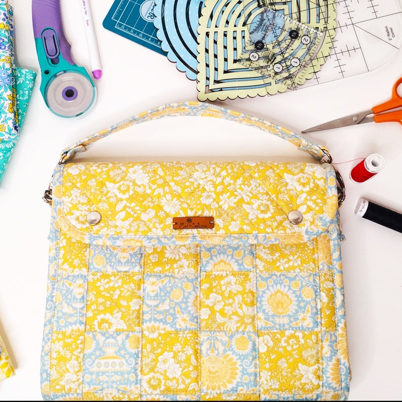 May include: A yellow and blue floral fabric handbag with a flap closure and a top handle. The bag has a quilted design and a small brown leather tag with the text "The Maker's Workshop" on it.