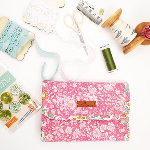 May include: A pink floral fabric wallet with a brown leather tag that says "Art Gallery Fabrics". The wallet is surrounded by various craft supplies, including lace, thread, and scissors.
