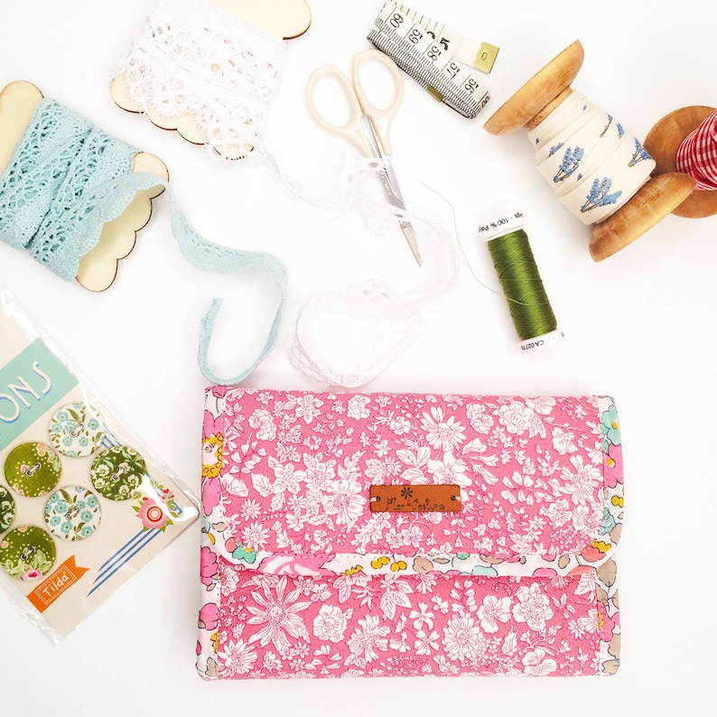 May include: A pink floral fabric wallet with a brown leather tag that says "Art Gallery Fabrics". The wallet is surrounded by various craft supplies, including lace, thread, and scissors.