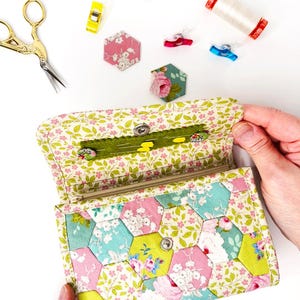 May include: A floral patterned fabric wallet with a zipper closure and a snap button. The wallet is open and the inside is lined with a green fabric. The wallet is made of patchwork fabric with a hexagonal pattern.