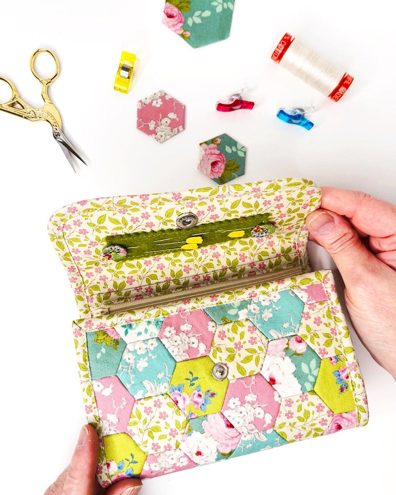 May include: A floral patterned fabric wallet with a zipper closure and a snap button. The wallet is open and the inside is lined with a green fabric. The wallet is made of patchwork fabric with a hexagonal pattern.