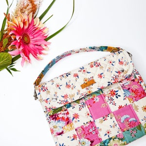 May include: A white quilted crossbody bag with a patchwork design featuring pink, blue, and green floral fabric. The bag has a flap closure and a small brown leather label.