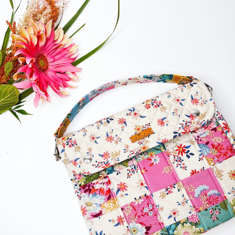 May include: A white quilted crossbody bag with a patchwork design featuring pink, blue, and green floral fabric. The bag has a flap closure and a small brown leather label.