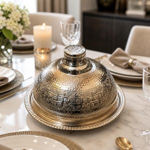 Peut inclure: Un plat de service argenté avec un couvercle en forme de dôme, présentant une texture martelée et des motifs en relief complexes. Le plat repose sur un plateau rond à bord perlé. L'ensemble comprend des assiettes blanches, de la verrerie et une bougie allumée.