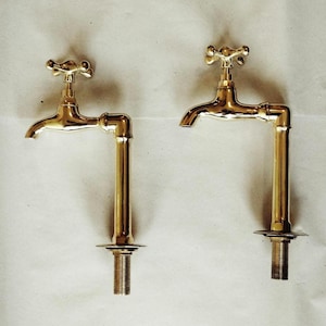 K&ouml;nnte beinhalten: Zwei goldfarbene Wasserh&auml;hne mit kreuzf&ouml;rmigen Griffen und einer rohrartigen Verl&auml;ngerung. Die Wasserh&auml;hne sind auf einer wei&szlig;en Oberfl&auml;che montiert.