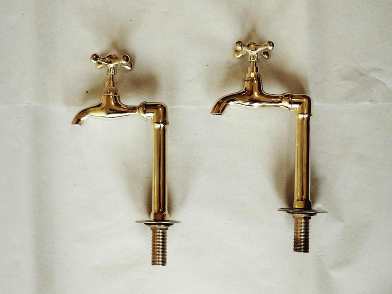 K&ouml;nnte beinhalten: Zwei goldfarbene Wasserh&auml;hne mit kreuzf&ouml;rmigen Griffen und einer rohrartigen Verl&auml;ngerung. Die Wasserh&auml;hne sind auf einer wei&szlig;en Oberfl&auml;che montiert.