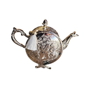 Peut inclure: Une théière en argent avec un motif orné et un pompon blanc. La théière a un corps rond et un bec verseur sur le côté. La théière est posée sur un fond blanc.