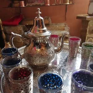 Peut inclure: Une théière marocaine en argent avec des motifs complexes est posée sur un plateau en argent avec six verres. Les verres sont remplis d'un liquide foncé et présentent des motifs complexes. Les verres sont dans des tons de bleu, violet et rouge.