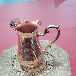 Könnte beinhalten: Eine gehämmerte Kupferkanne mit einer breiten Öffnung und einem gebogenen Griff. Die Kanne steht auf einem dekorativen Tablett mit einem komplizierten Design.