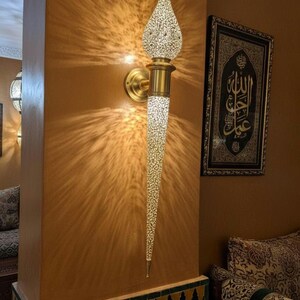 May include: A brass wall sconce with a textured, elongated design. The sconce has a teardrop-shaped top and a long, tapered body. The light casts a pattern on the wall. A framed artwork hangs nearby.