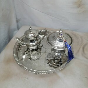 Peut inclure: Un plateau de service en argent avec un motif orné, comprenant une théière en argent et un sucrier en argent avec un pompon bleu.