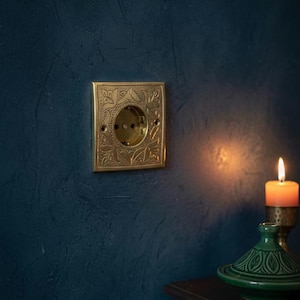 May include: A brass electrical outlet with an ornate floral design is mounted on a dark blue wall. The outlet has a square faceplate with a circular opening for the plug. A lit candle and a green decorative piece are in the background.