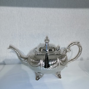 Peut inclure: Une théière argentée avec des détails décoratifs. Elle présente un bec verseur incurvé, une anse en boucle et un couvercle avec un bouton décoratif. Elle repose sur trois petits pieds et possède un pompon noir.