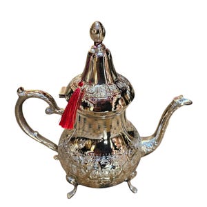 Peut inclure: Une théière en argent avec un design orné et un pompon rouge. La théière a un corps rond et un couvercle haut et bombé. Elle repose sur trois pieds.